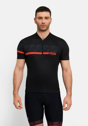 Mann trägt schwarzes Radtrikot mit rotem Streifen und schwarze Radhose mit rotem Muster, steht vor einem einfarbigen Hintergrund.