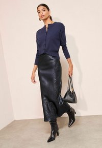 Cardigan închis la culoare cu nasturi și fustă maxi din satin negru lucios cu o siluetă ajustată. Botine negre cu vârf ascuțit până la gleznă și o geantă mică neagră.