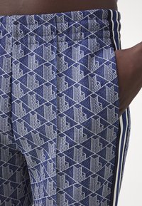 Close-up of blue pants with white geometric pattern and vertical navy, white, and black side stripes, hand resting in pocket.
