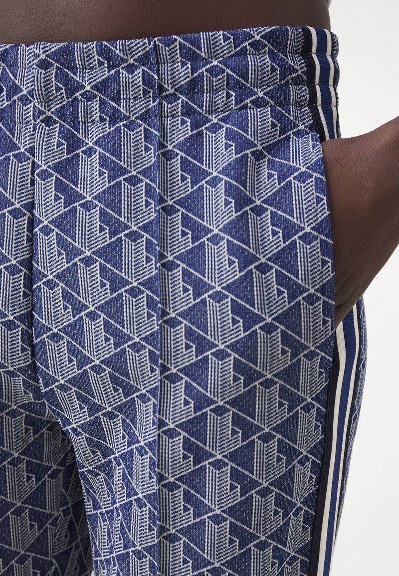 Zbliżenie na niebieskie spodnie z białym geometrycznym wzorem oraz pionowymi paskami w kolorze granatowym, białym i czarnym, ręka spoczywająca w kieszeni.