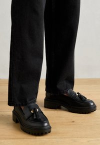 Mustad nahast loafers, millel on paks tald ja dekoratiivne tuttu ning kuldne detail. Kannab tumedate teksadega puidust pinnal.