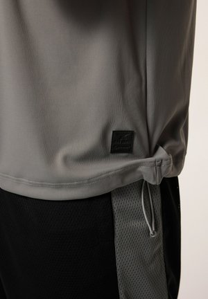 Maglietta atletica grigia con tessuto liscio, dotata di una toppa logo nera, cordino laterale e abbinata a pantaloni corti in rete neri.