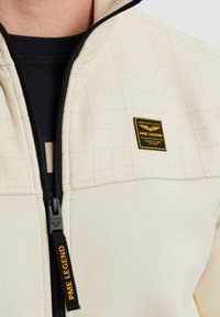 Veste zippée blanc cassé avec des sections supérieures matelassées, fermeture éclair noire et un label PME Legend jaune sur la poitrine. Tissu doux avec une finition texturée.