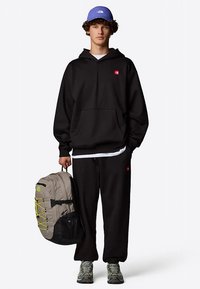 Černá mikina s kapucí a tepláky s červeným logem, doplněné světle šedým batohem se žlutými detaily a modrou čepicí.