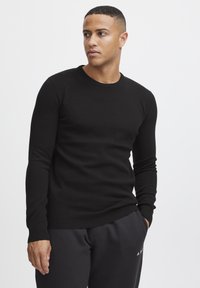 Black knit sweater with textured pattern, round neckline, and long sleeves. Worn with dark sweatpants, featuring a ribbed hem and cuffs.