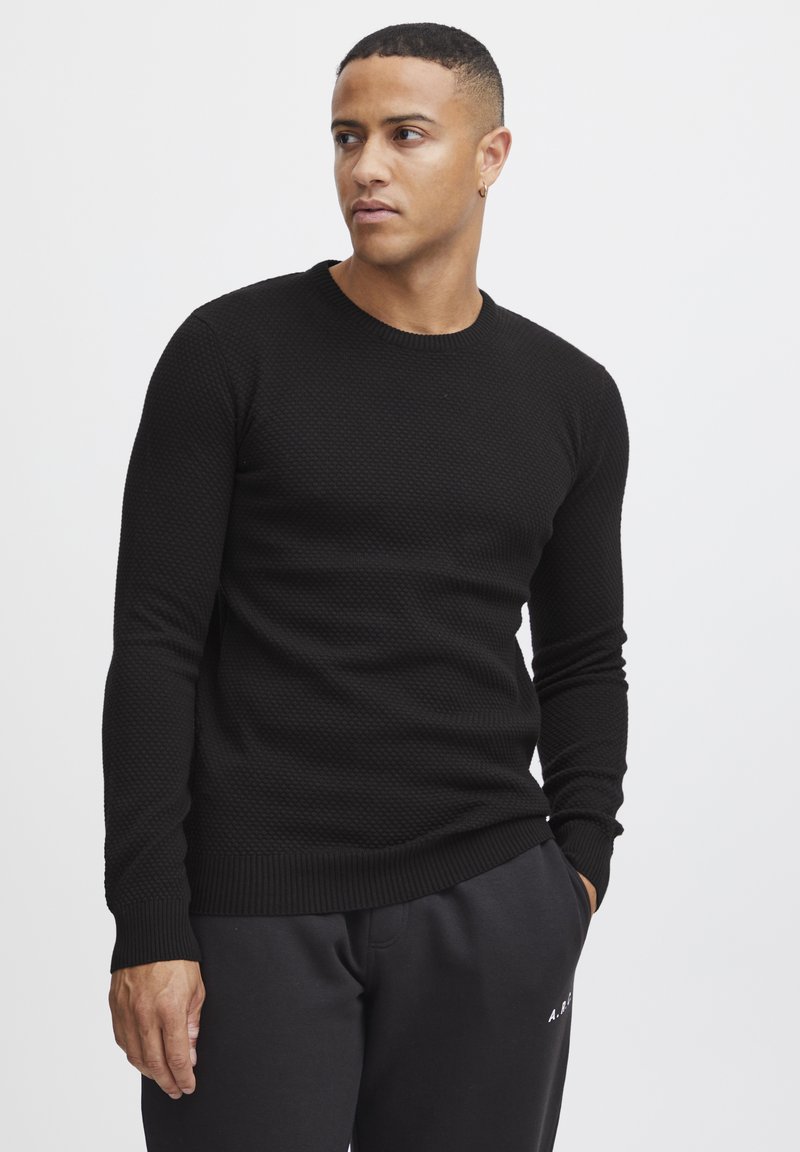 Black knit sweater with textured pattern, round neckline, and long sleeves. Worn with dark sweatpants, featuring a ribbed hem and cuffs.