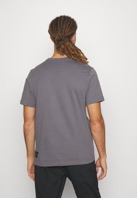 Muž s dredy svázanými dozadu, oblečený v jednoduchém šedém tričku s krátkým rukávem a černých kalhotách, otočený zády proti bílému pozadí.