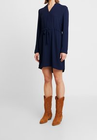 Robe à manches longues bleu marine avec un col en V et une ceinture ajustable à la taille, associée à des bottes de cowboy en daim beige avec des détails brodés.