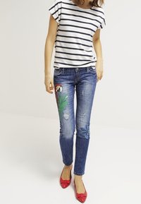 Camisa a rayas de manga corta combinada con jeans de mezclilla azul con diseños bordados de loro y flores. Modelo lleva zapatos planos rojos.