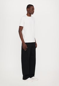 T-shirt en coton blanc à manches courtes, assortie à un pantalon large noir et des baskets blanches, sur fond uni.