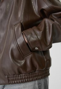 Poignet d'une veste en cuir marron avec un bouton pression et main partiellement à l'intérieur de la poche latérale, portée sur un jean en denim gris clair.
