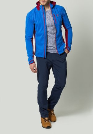 Mann in blau-roter Zip-Jacke, grauem Shirt, dunkler Hose und beige Wanderstiefeln, steht mit einer Hand in der Tasche.