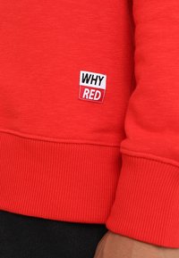 Manga y dobladillo de sudadera roja con un pequeño parche blanco y rojo que muestra el texto "¿POR QUÉ ROJO?" en la parte delantera inferior.