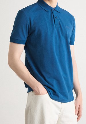 Man die een blauw poloshirt met korte mouwen draagt met het logo "BOSS" en een witte broek, staand met één hand in de zak tegen een effen achtergrond.