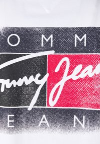 Mustavalkoinen ja punainen lohkomuotoilu tyylitellyllä valkoisella tekstillä "Tommy Jeans" sekä "TOMMY JEANS" suurilla kirjaimilla kankaalla.