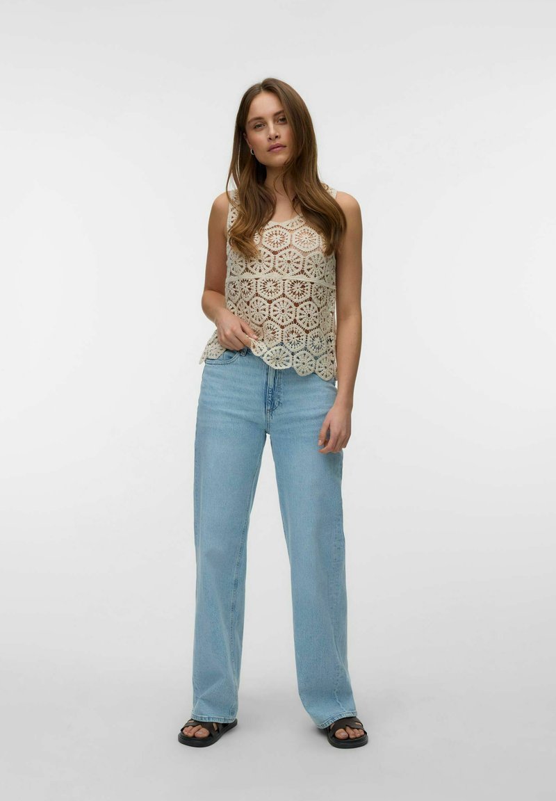Jeune femme debout portant un haut beige en crochet sans manches, un jean bleu clair à jambes larges et des sandales noires sur fond blanc uni.