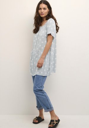 Tunique bleu clair avec motif floral blanc, manches courtes et coupe décontractée, associée à un jean en denim retroussé et des sandales noires.