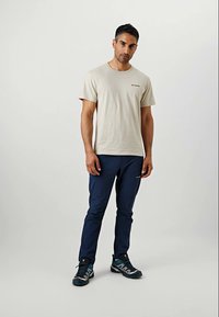 Beige cotton t-shirt with short sleeves and embroidered logo, paired with navy stretch pants and gray and teal athletic shoes.