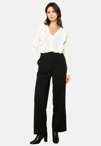 White ruffled blouse with a V-neck, paired with black high-waisted trousers. Model wearing black heeled ankle boots.