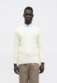 Pehmeä kermaisen värinen neule, jossa on pyöreä pääntie, yhdistetty vaaleansiniseen kauluspaitaan ja ruskeisiin, kuvioituihin housuihin. Ei näkyviä logoja tai yksityiskohtia.