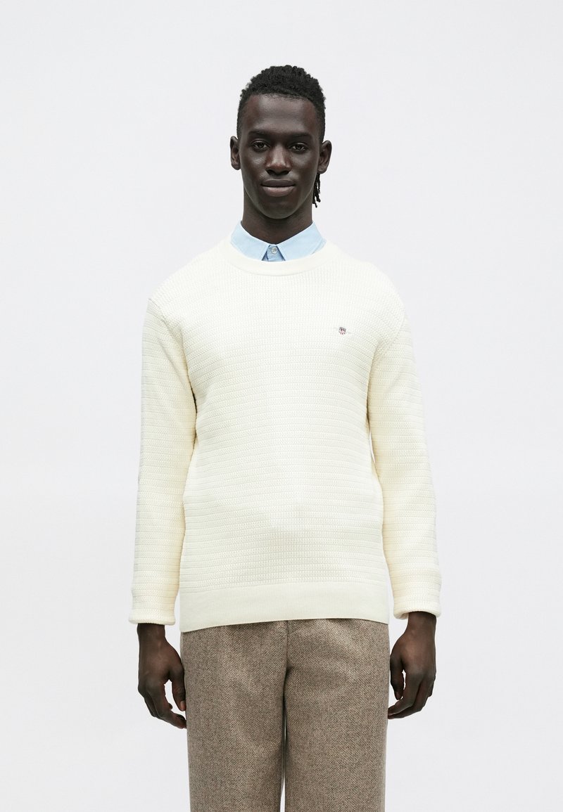 Pehmeä kermaisen värinen neule, jossa on pyöreä pääntie, yhdistetty vaaleansiniseen kauluspaitaan ja ruskeisiin, kuvioituihin housuihin. Ei näkyviä logoja tai yksityiskohtia.