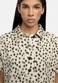 Koszulka z krótkim rękawem, z kołnierzykiem, wykonana z beżowego materiału z czarnym wzorem w kropki i czterema czarnymi guzikami z przodu.