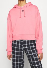 Osoba w różowej bluzie z kapturem Adidas z troczkami oraz czarno-białych spodniach w kratę, stojąca na tle jednolitego tła.