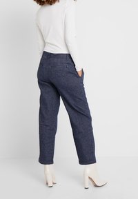 Donkerblauwe, licht taps toelopende broek met opgestikte zakken, gemaakt van textuurstof; gecombineerd met een licht T-shirt met lange mouwen en beige hoge hakken.