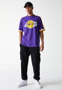 Lila T-shirt med korta ärmar och gula accenter samt tryck med texten "Los Angeles Lakers". Matchad med svarta cargobyxor och vita sneakers.