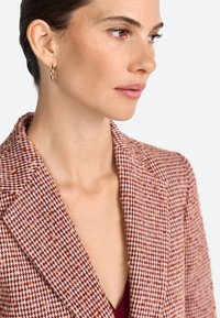 Blazer houndstooth bertekstur, berwarna merah muda dan marun dengan potongan yang dirancang dan lapel bergerigi, dipakai di atas atasan burgundy. Anting-anting hoop emas sebagai aksen.