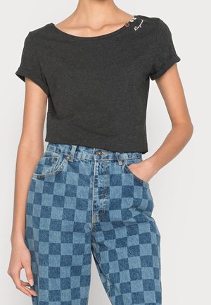 Mujer con camiseta de manga corta gris oscuro con botones decorativos y vaqueros azules a cuadros de talle alto, una mano en el bolsillo.