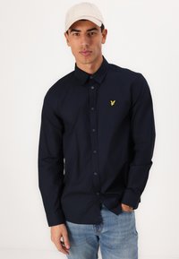 Cămașă cu nasturi de marină, cu mâneci lungi și un logo galben brodat. Modelul poartă blugi albaștri deschiși și o pălărie de culoare deschisă.