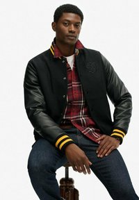Zwart varsityjack met leren mouwen, gele gestreepte manchetten en geborduurd logo. Draag het over een rood geruit overhemd en donkere jeans.