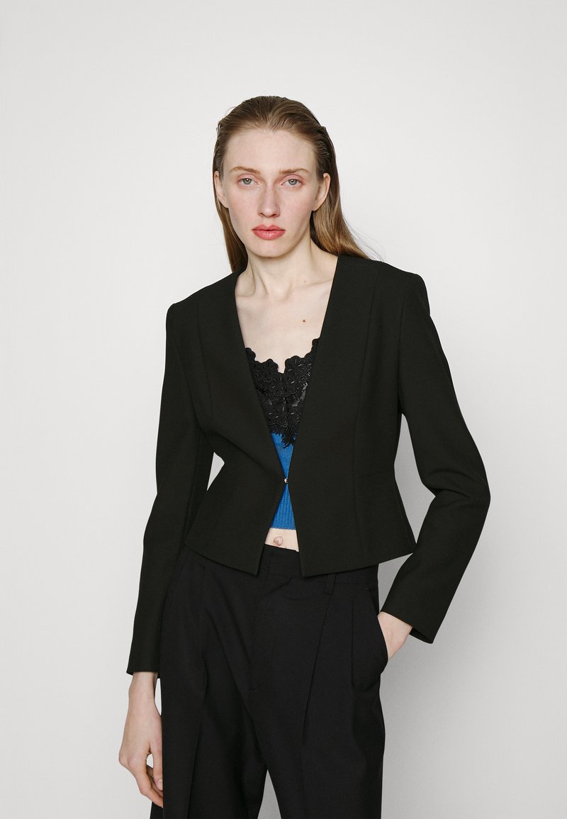 Svart kort blazer med en strukturert design og lange ermer, kombinert med en blå ribbet topp med svarte blonder.