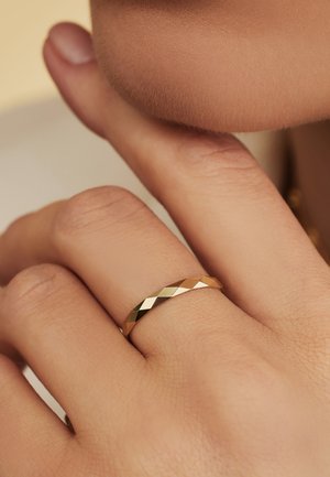 Goldfarbener Bandring mit facettiertem Design. Die glatte Textur und die eckigen Kanten erzeugen ein geometrisches Muster. Am Hand mit natürlichem Hautton getragen.