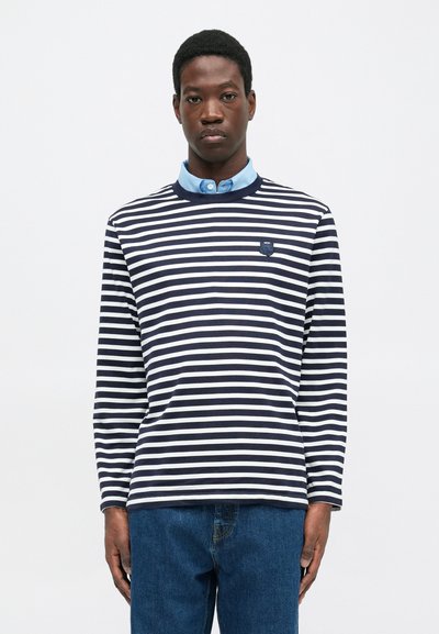 Long-sleeve striped shirt in navy and white cotton, featuring a round neckline and a small embroidered logo on the chest, layered over a blue collared shirt.
