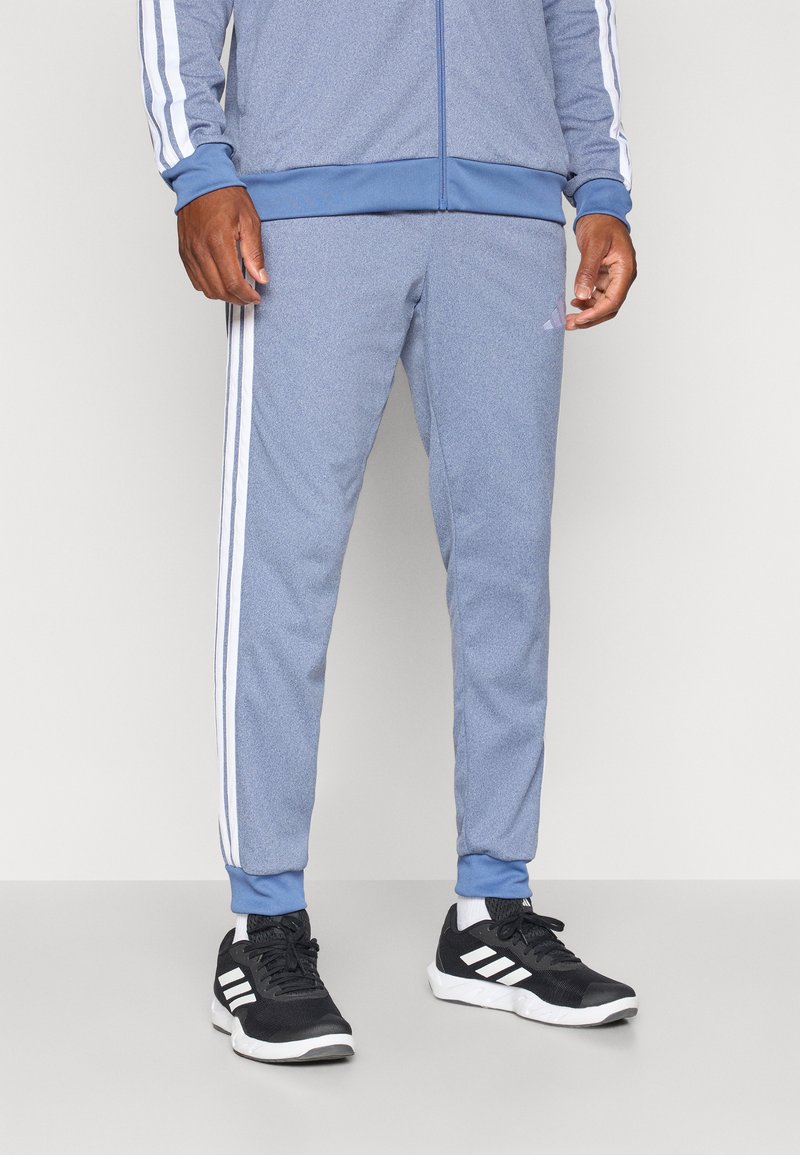 Pantalons de sport bleu clair en tissu texturé, avec des rayures blanches sur les côtés, des poignets côtelés et un logo brodé sur la jambe.