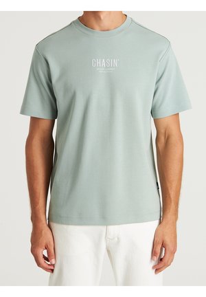 Homme portant un t-shirt vert clair à manches courtes avec le texte "CHASIN'" et un pantalon blanc, montré de la nuque aux hanches, debout devant un fond uni.
