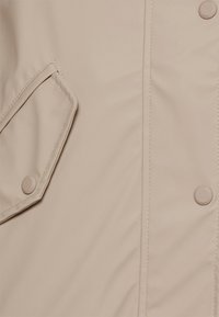 Lichte beige regenjas met een soepele textuur, drukknoopsluiting en een diagonale klepzak aan de voorkant aan de linkerkant.