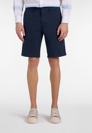 Uomo che indossa pantaloncini blu navy fino al ginocchio, camicia bianca con bottoni e sneakers beige, in piedi con le mani rilassate ai lati.
