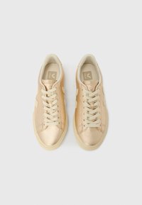 Par metalliske gullfargede sneakers med snøring, beige såler og Veja-logo på innsiden, sett ovenfra på en lys bakgrunn.