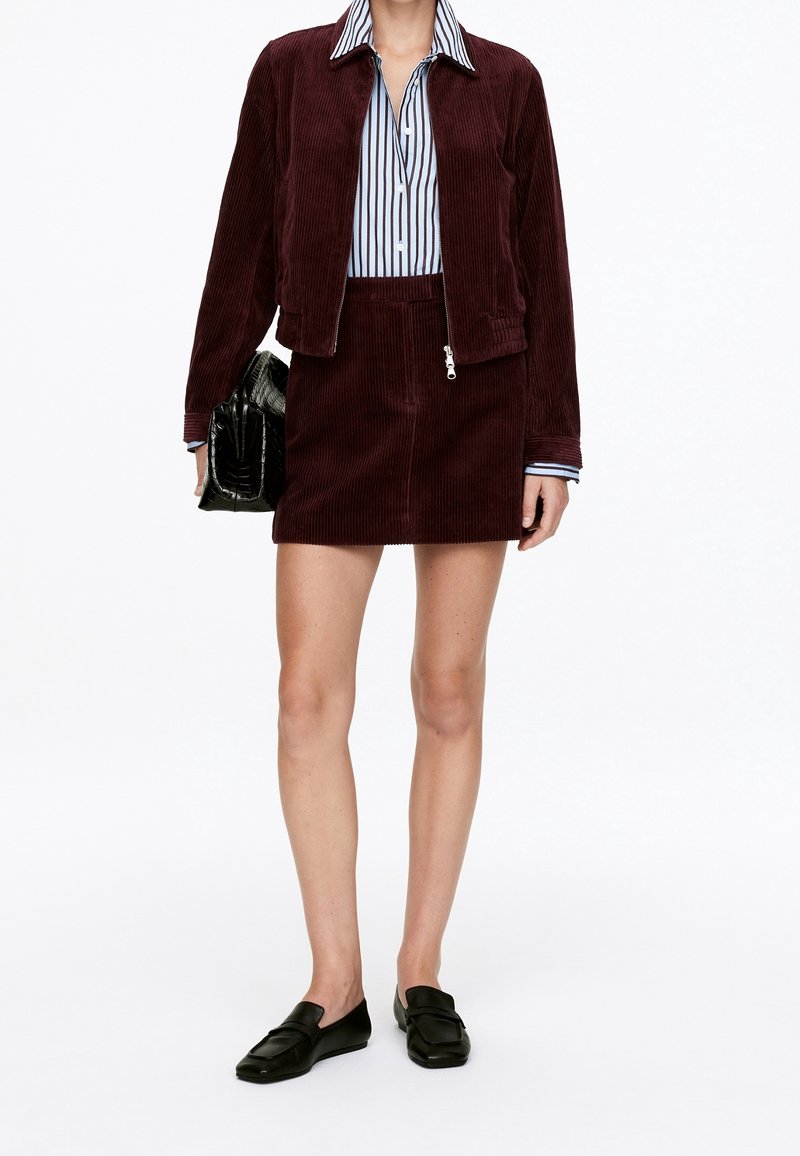 Femme portant une veste et une jupe en velours côtelé bordeaux avec une chemise rayée boutonnée, tenant un sac à main noir et portant des mocassins noirs.