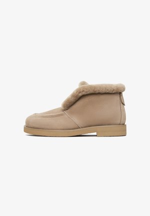 Stivale alla caviglia in suede beige con un soffice collo in shearling, punta tondeggiante e suola piatta bassa. Design semplice con una cerniera laterale per un facile accesso.
