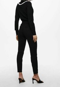 Haut noir à manches longues avec un col à volants blancs, associé à un pantalon noir ajusté et des sandales à talons noires. Tissu lisse, silhouette élancée.