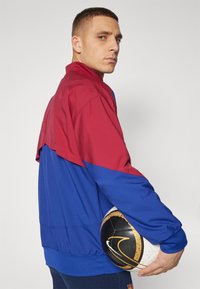 Färgglad jacka i röd och blå tyg, med hög krage och elastiska ärmslut. En svart och guldig basketboll hålls under armen.