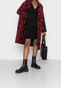 Rot-schwarz gemusterter Mantel über einem schwarzen, gerippten Kleid, kombiniert mit klobigen schwarzen Stiefeletten und einer schwarzen Handtasche.