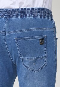 Jeans de denim con una cinturilla elástica de color azul oscuro, lavado azul claro y un bolsillo trasero con una etiqueta cuadrada y costuras decorativas.