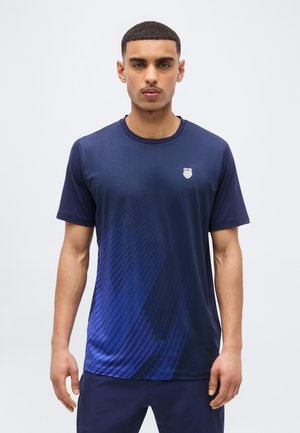 Młody mężczyzna ubrany w granatowy sportowy t-shirt z ukośnymi wzorami i małym logo na lewej piersi, stojący na jednolitym tle.