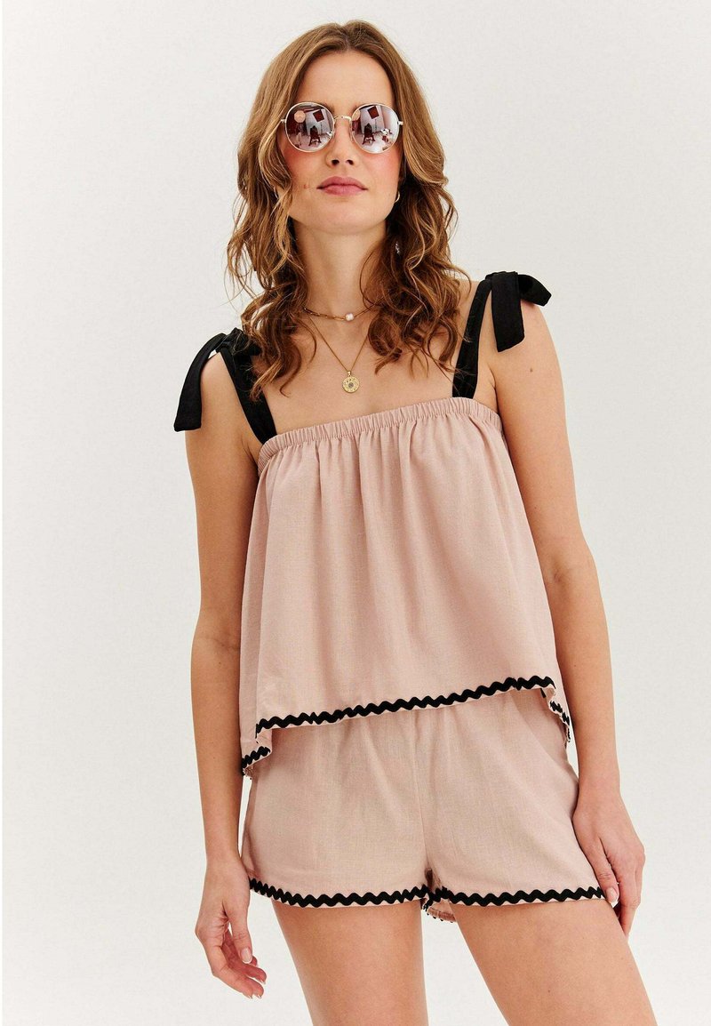 Žena v svetloružovom dvojdielnom oblečení s čiernym lemom, čiernymi ramennými viazaniami, okrúhlymi slnečnými okuliarmi a viacvrstvovými zlatými náhrdelníkmi na bielom pozadí.