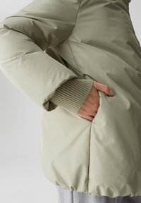 Giacca imbottita verde chiaro con una texture liscia, polsini a costine e tasca laterale, caratterizzata da un orlo inferiore arricciato per una vestibilità aderente.